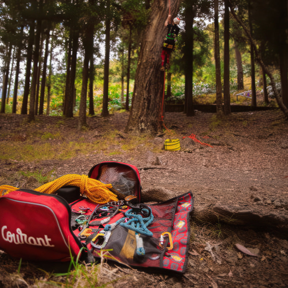 A red Courant climbing bag filled with climbing gear, including a yellow rope and carabiners, set on a forest floor with a climber visible high up a tree in the background. Available in 54L & 75L