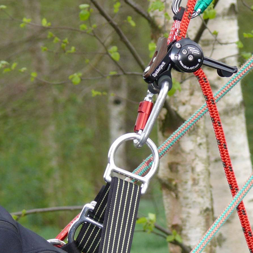 Close-up of a climbing harness and ART Spiderjack 2.1 Dyneema, in use with red and green ropes, a black harness strap, and metal hardware against a blurred tree background.